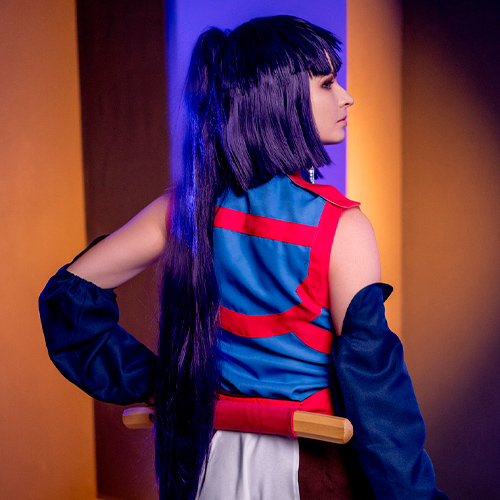 Akatsuki cosplay from Log Horizon. The model is standing with her back to the camera, showcasing her long purple hair tied in a ponytail. She is holding a kunai in her right hand and a sword is sheathed on her back. The ninja outfit is accented by blue and red, and the background is bathed in contrasting blue and orange lighting.