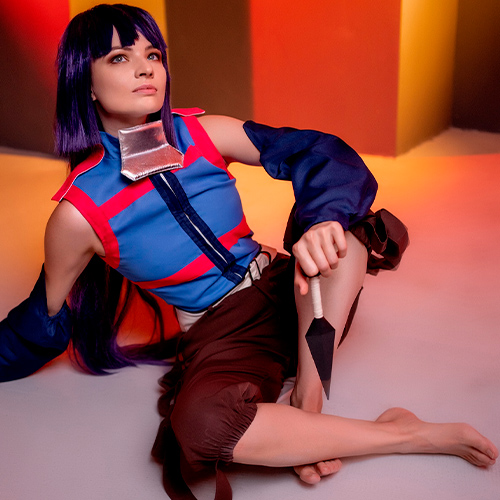 Akatsuki cosplay from Log Horizon. The model is sitting on the ground in a relaxed yet poised manner, holding a kunai in her right hand. She wears a blue and red ninja outfit, and her expression is thoughtful as she gazes into the distance. The background features warm tones of red and orange, contrasting with the cool blue of her costume.