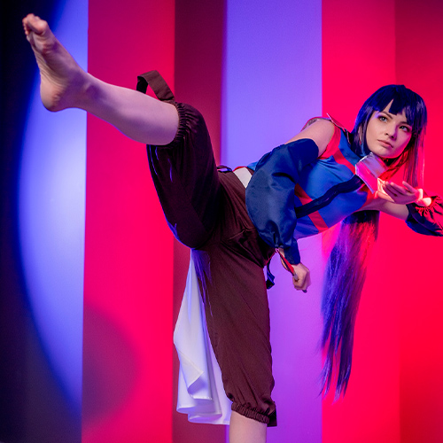 Akatsuki cosplay from Log Horizon. The model executes a high kick, demonstrating a powerful martial arts move. She wears a determined expression and her vibrant blue and red ninja outfit contrasts against the dramatic background lighting of red and blue.