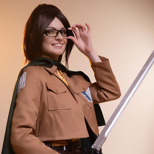 Hanji Zoe cosplay from Attack on Titan, seated on a stool with a Survey Corps uniform, glasses, green cape, and sword, smiling confidently while adjusting her glasses in warm studio lighting