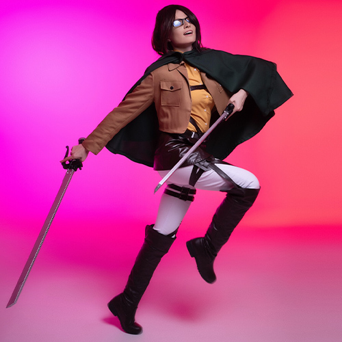 Dynamic Hanji Zoe cosplay from Attack on Titan, striking a mid-action pose with dual blades and a green cape flowing, set against a vibrant pink and red gradient background