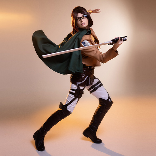 Hanji Zoe cosplay from Attack on Titan, captured mid-swing with a blade and green cape in motion, set against a soft beige backdrop with warm lighting