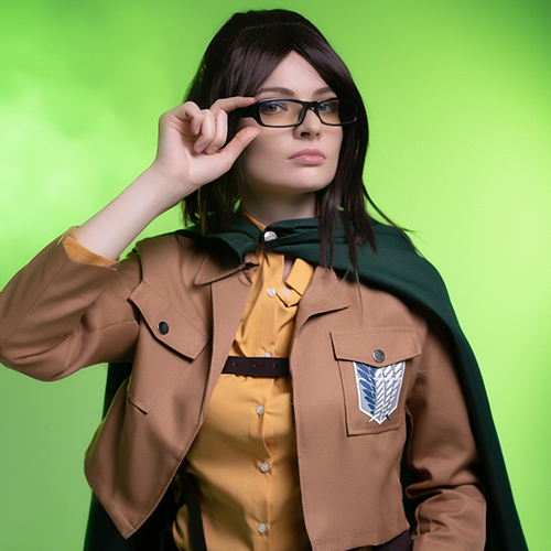 Hanji Zoe cosplay from Attack on Titan, character confidently adjusting glasses with a focused expression, set against a vivid green background with dramatic lighting