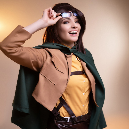 Hanji Zoe from Attack on Titan cosplay with a joyful expression, playfully lifting her glasses, in full Survey Corps uniform, highlighted by warm lighting