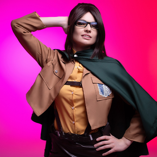 Hanji Zoe cosplay from Attack on Titan, posing confidently in Survey Corps uniform with a vibrant pink backdrop, highlighting her glasses and green cape