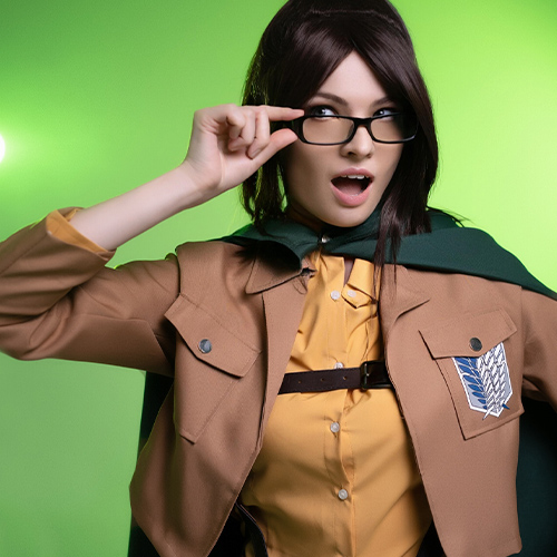 Hanji Zoe cosplay from Attack on Titan, making a playful expression while adjusting her glasses, wearing the classic Survey Corps uniform with a green cape, standing against a bright green background