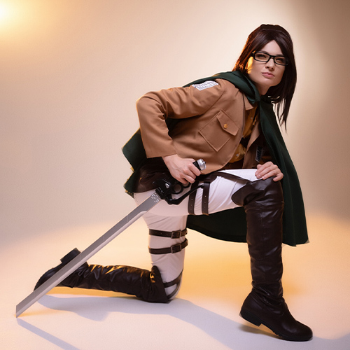 Hanji Zoe cosplay from Attack on Titan, kneeling with a determined look, holding a sword in the iconic Survey Corps uniform with a green cape, staged under a warm studio light