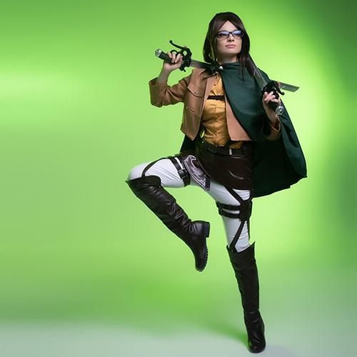 Hanji Zoe cosplay from Attack on Titan, in a dynamic pose holding dual blades, wearing the full Survey Corps uniform with green cape, glasses on, against a bright green studio background