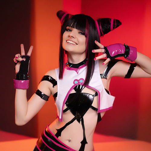 Cosplayer dressed as Juri Han from Street Fighter, striking a playful pose with both hands showing peace signs, smiling confidently against a warm red and orange backdrop