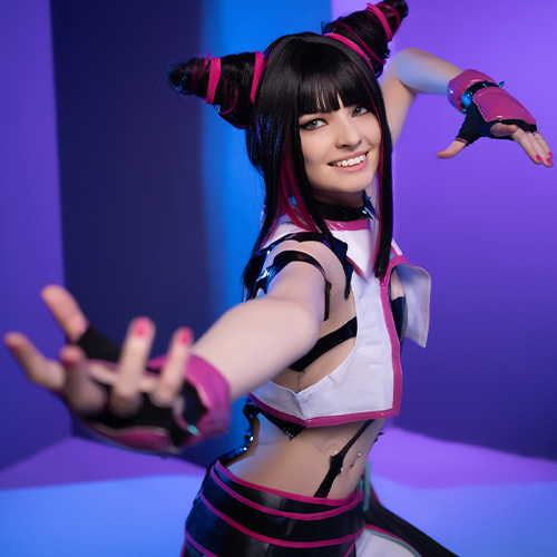 Cosplayer portraying Juri Han from Street Fighter, striking a dynamic pose with an extended hand and a confident smile. The costume features pink and black elements, with her hair styled in two high buns wrapped in pink ribbons, set against a vibrant blue and purple background