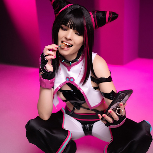 Cosplayer dressed as Juri Han from Street Fighter, squatting while holding a phone in one hand and a lollipop in the other. Her black and pink outfit is accentuated by the vibrant pink lighting in the background, capturing her playful and rebellious persona