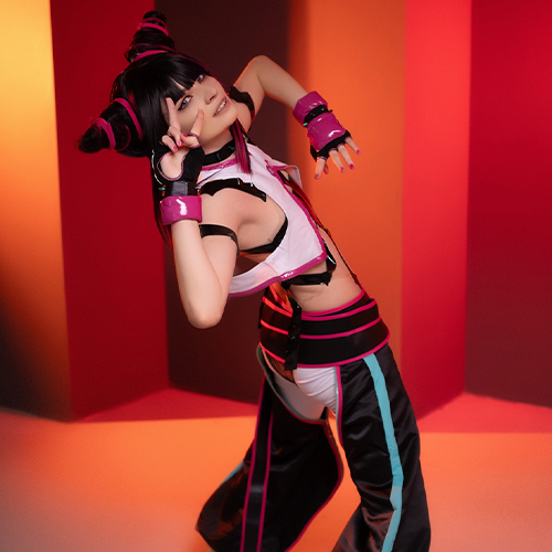 Cosplayer dressed as Juri Han from Street Fighter, striking a playful pose with a peace sign. The lighting shifts from warm orange to red, complementing her energetic stance and vibrant outfit, featuring black, pink, and white colors. The background enhances the dynamic mood of the image