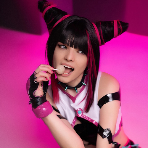 Cosplayer dressed as Juri Han from Street Fighter, sitting on the floor while holding a lollipop. The vibrant pink and purple tones in her outfit and hairstyle are accentuated by the neon pink lighting in the background, creating a playful and energetic atmosphere