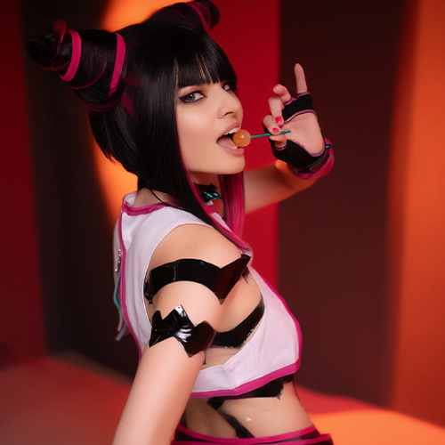 Cosplayer portraying Juri Han from Street Fighter, in a playful pose while holding a lollipop. The character's iconic hairstyle, with twisted buns wrapped in pink ribbons, is striking. The vivid orange background complements the pink accents on her outfit, highlighting the mischievous and bold personality of the character