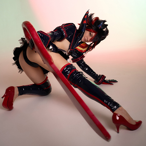 Cosplayer in an action-ready pose as Ryuko Matoi from Kill la Kill. Wearing a black and red latex version of the Senketsu Kamui, the cosplayer wields Ryuko's iconic oversized red scissor blade with precision and strength. Her crouched stance and intense expression bring out the dynamic energy of the character. The combination of red heels and dramatic lighting enhance the fierce and bold aesthetic of the costume