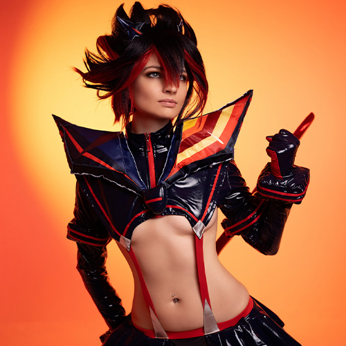 Fierce Ryuko Matoi cosplay striking a powerful pose, with the orange backdrop amplifying the intensity of her red and black outfit from Kill la Kill. The intricate detailing of the costume is on full display, radiating strength and attitude