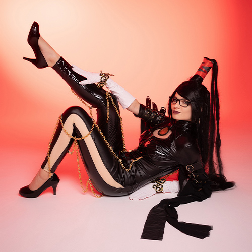 Stylish Bayonetta cosplay striking a bold and playful pose, showcasing the character's signature long black hair and form-fitting black outfit adorned with gold chains. The vibrant red background enhances the character's confidence and flair