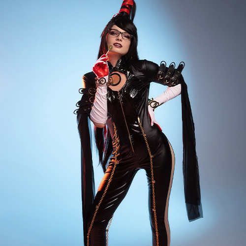 Confident Bayonetta cosplay, showcasing the character’s iconic tight black suit adorned with gold chains and flowing sleeves. The cosplayer adds flair with a red glove and a playful pose, set against a soft blue backdrop that contrasts with her dark, edgy outfit