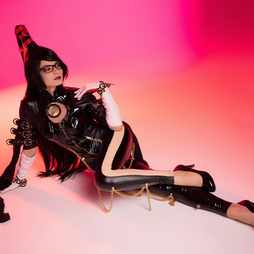 Striking Bayonetta cosplay reclining against a gradient pink and orange background. The glossy black bodysuit, complete with gold chains and accessories, is complemented by her signature glasses and black heels, embodying the character’s fierce and seductive style