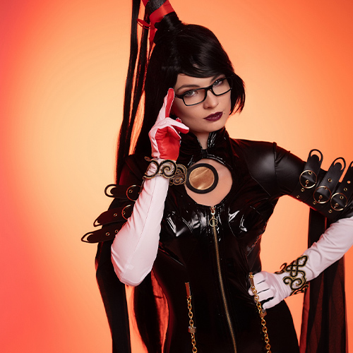 Bayonetta cosplay strikes a poised pose with a serious expression against a bold orange backdrop. The character’s iconic black latex suit with intricate gold details is enhanced by the signature long hair styled into a high ponytail, complemented by her sharp glasses and red gloves