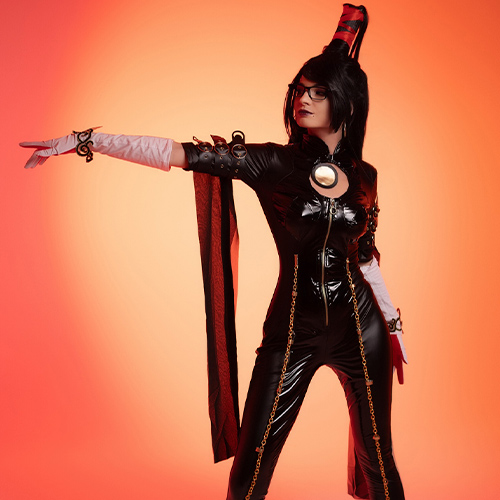 Striking pose of Bayonetta cosplay in a black latex suit with gold chains and a high ponytail, extending a gloved hand forward. The scene is set against a bold orange backdrop, emphasizing the dynamic and confident stance