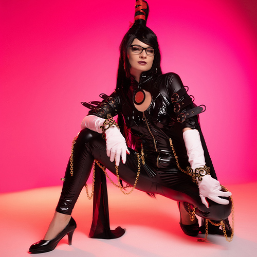 Bayonetta cosplay crouching in a powerful pose, wearing a black latex bodysuit with gold chains, white gloves, and glasses. Set against a vibrant pink background, the character exudes confidence and style, with long black hair styled in a high ponytail