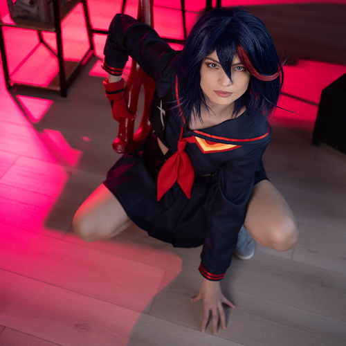 Dynamic Ryuko Matoi cosplay in action, crouching on the floor with her signature red Scissor Blade poised for battle. The scene is lit with intense pink and red hues that enhance the bold colors of her navy and red school uniform, capturing the character's fierce determination and readiness for action