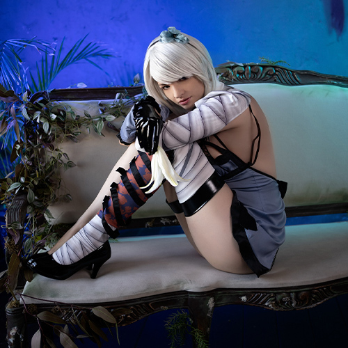 Kaine from Nier: Replicant sits curled on an elegant sofa, her posture reflective and guarded. The blue-toned background and lush greenery around her emphasize the melancholic beauty of her character, capturing both vulnerability and strength. Her bandaged arms and leg brace, along with her intense gaze, add depth to the scene, embodying Kaine’s resilience and complex personality. The soft lighting enhances the emotion, creating a serene yet haunting ambiance.