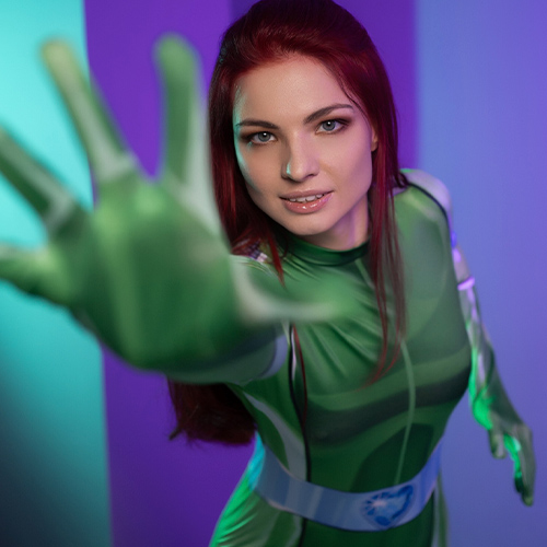 Dynamic Totally Spies Sam cosplay captured in an action pose, emphasizing her strong and determined nature. The vibrant green spy jumpsuit perfectly outlines the character’s sleek and powerful design, while her striking red hair adds an authentic touch to the look. With her hand extended dramatically towards the camera, this photo conveys energy and engagement, making it a stunning tribute to the iconic animated series.