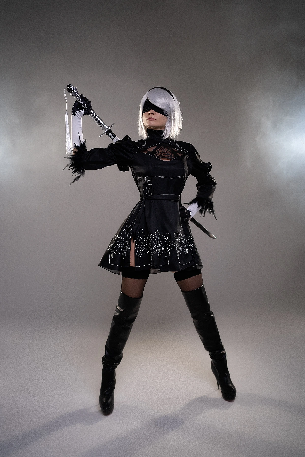 2B YoRHa cosplay standing with katana overhead, silver bob, black blindfold, embroidered dress, NieR Automata studio portrait