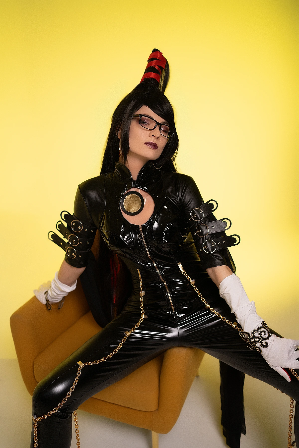 Bayonetta cosplay wide dominant chair pose by Neka-Chi, glossy black bodysuit with gold chain accessories, vivid yellow backdrop