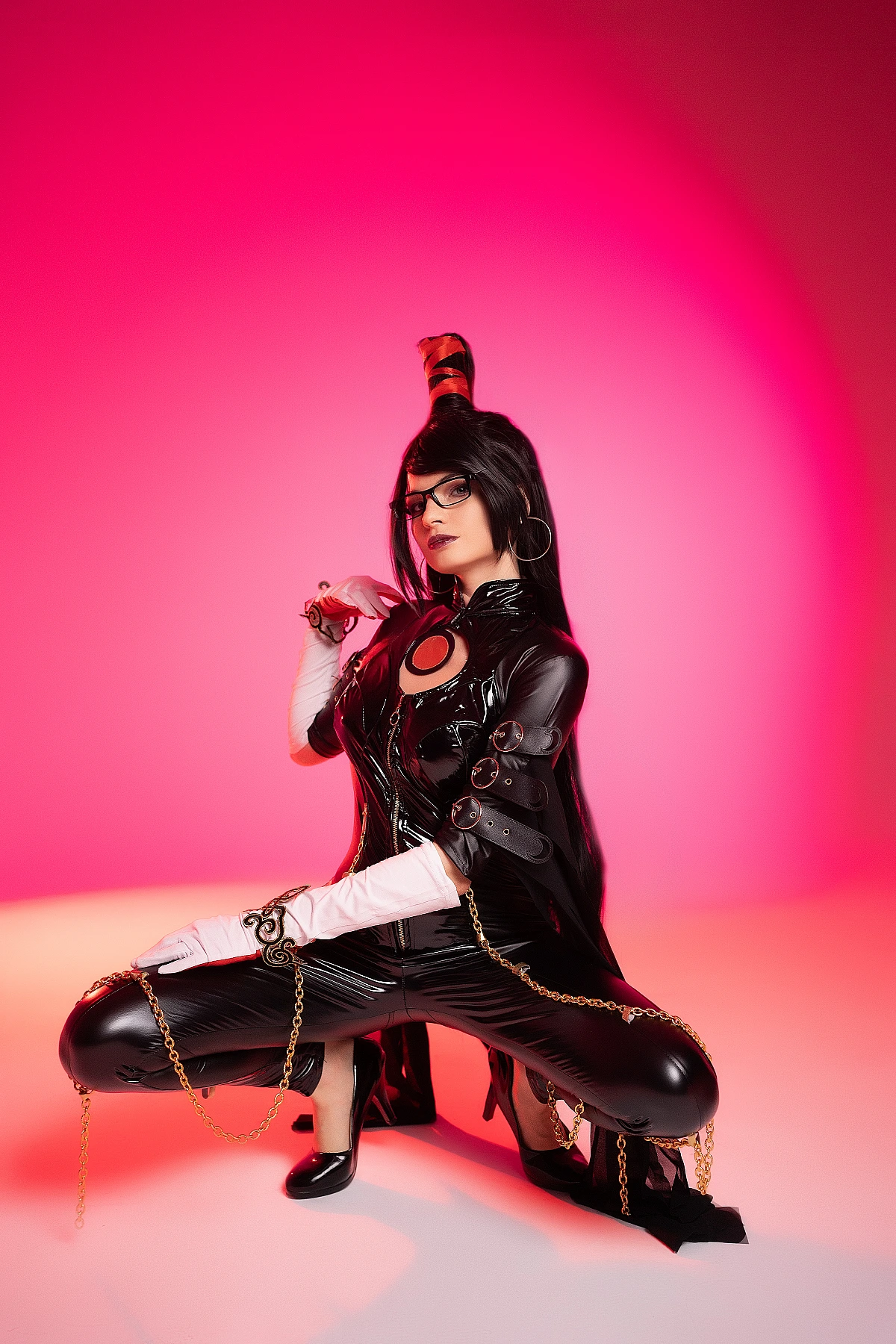 Bayonetta cosplay kneeling pose by Neka-Chi with white gloves, gold chains, glossy black bodysuit, pink-red gradient background
