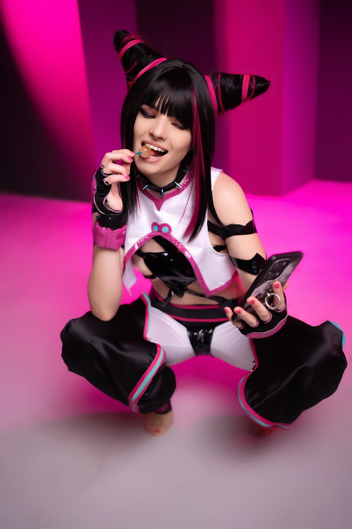 Juri Han cosplayer squatting with candy and smartphone, pink neon studio backdrop, glossy strap costume, horn buns, Street Fighter