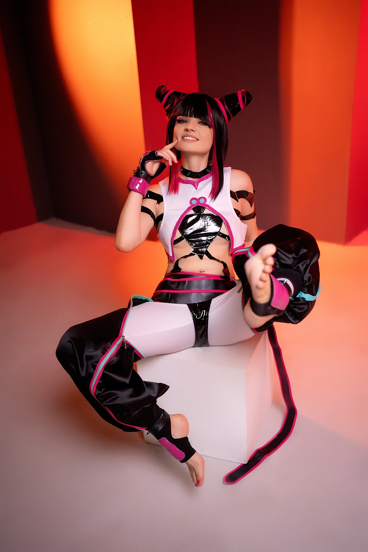 Juri Han cosplayer reclining on white cube with raised barefoot leg, red-orange neon striped backdrop, glossy strap costume, horn buns