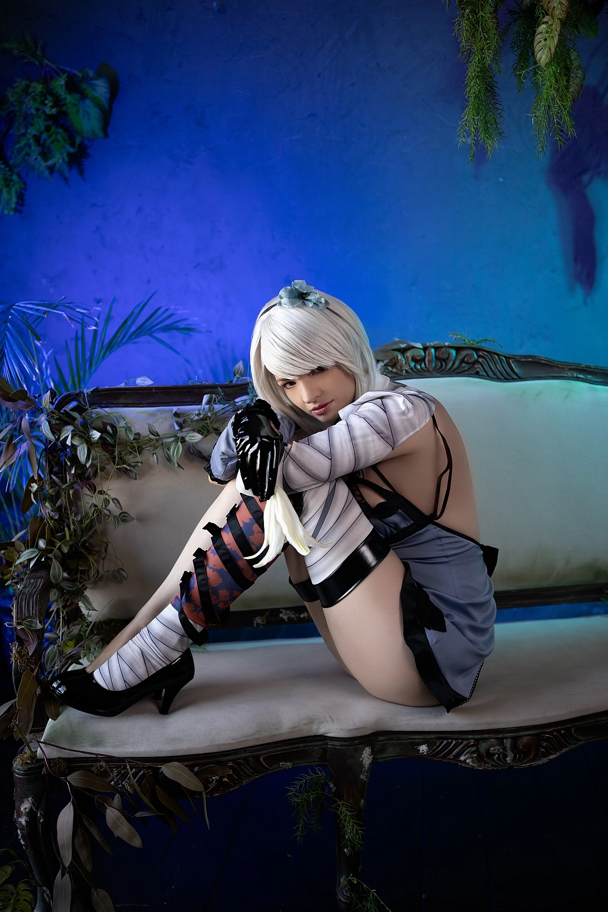 Kaine NieR Replicant cosplay curled introspective pose on sofa, silver bob hair, bandaged leg detail, deep blue background, soft shadows