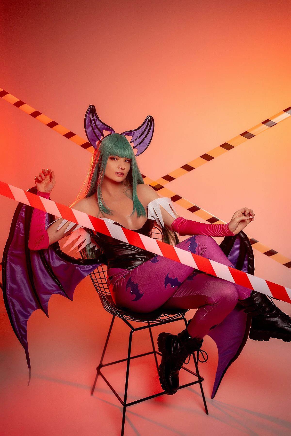 Seated Morrigan Aensland cosplay on wire chair, warm orange studio background, crossed tape lines, spread succubus wings
