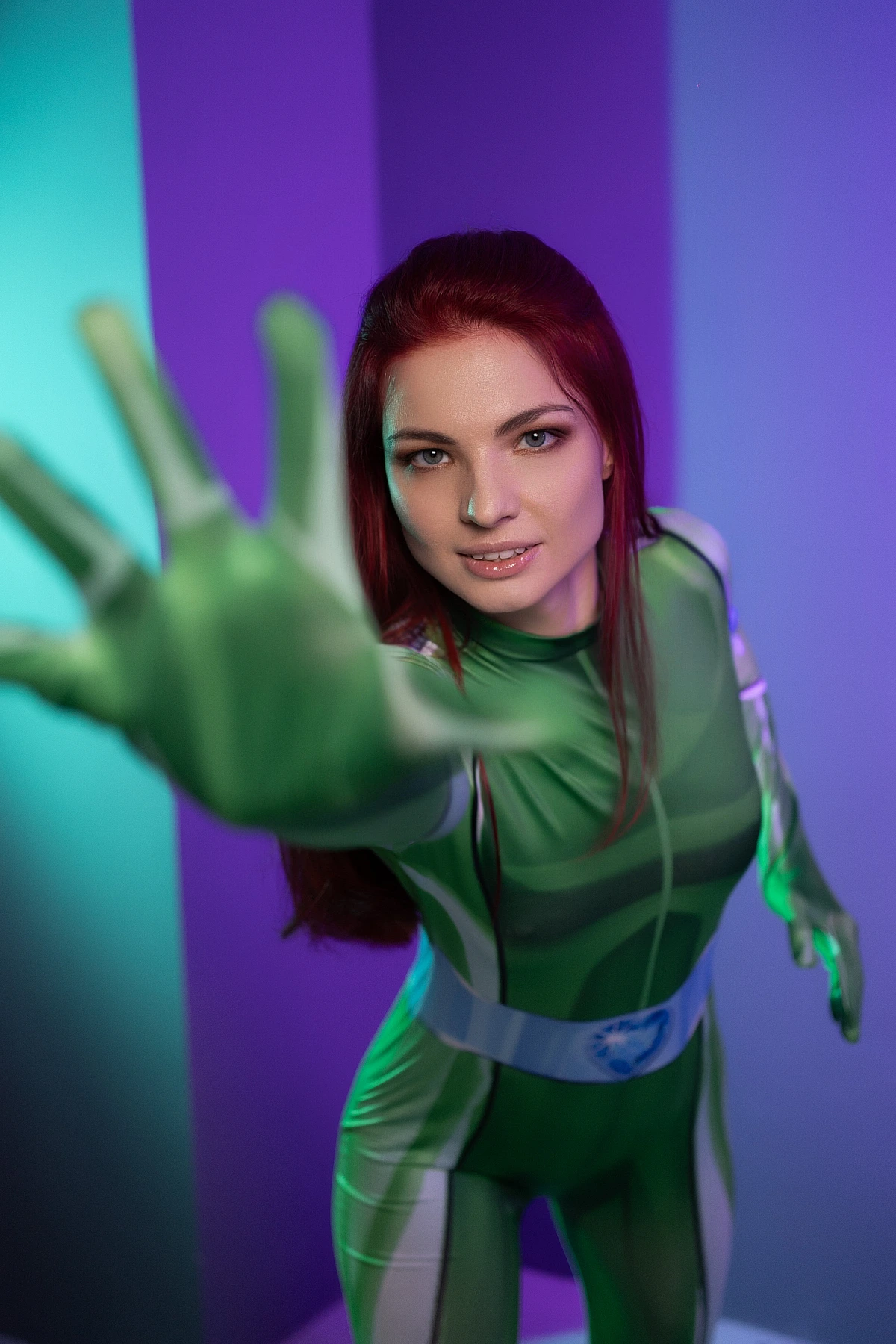 Dramatic perspective shot of Sam Totally Spies cosplay reaching toward camera in green catsuit with intense gaze and teal-violet studio lighting
