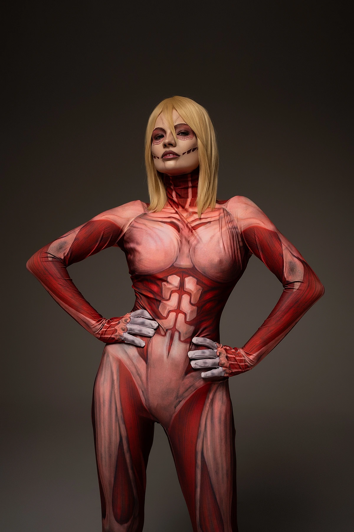 Female Titan cosplay hands on hips, Annie Leonhart dominance stance, anatomical muscle bodysuit, dark studio AOT cosplay