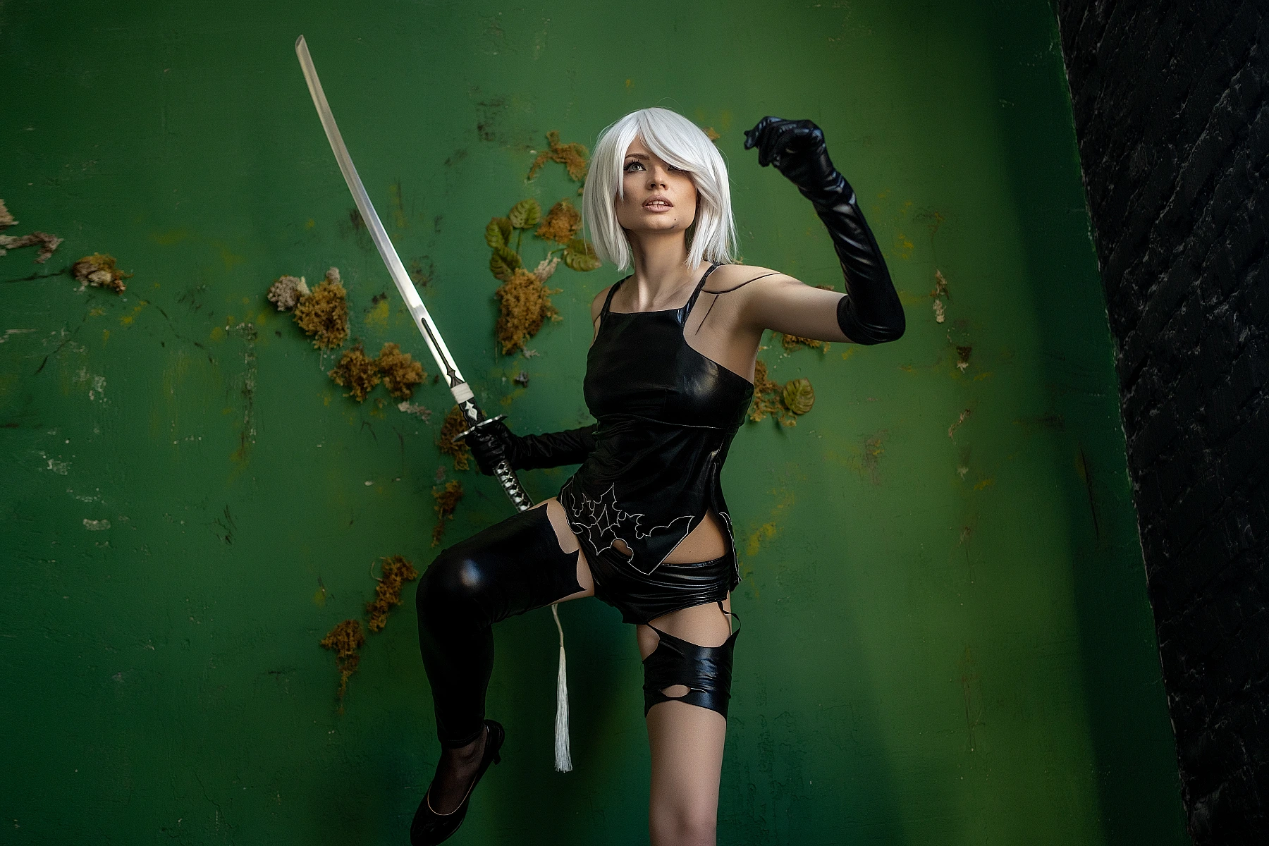 A2 NieR Automata cosplay action pose, raised katana sword, lifted leg dynamic stance, glossy black latex bodysuit, white bob hair, vivid green wall background