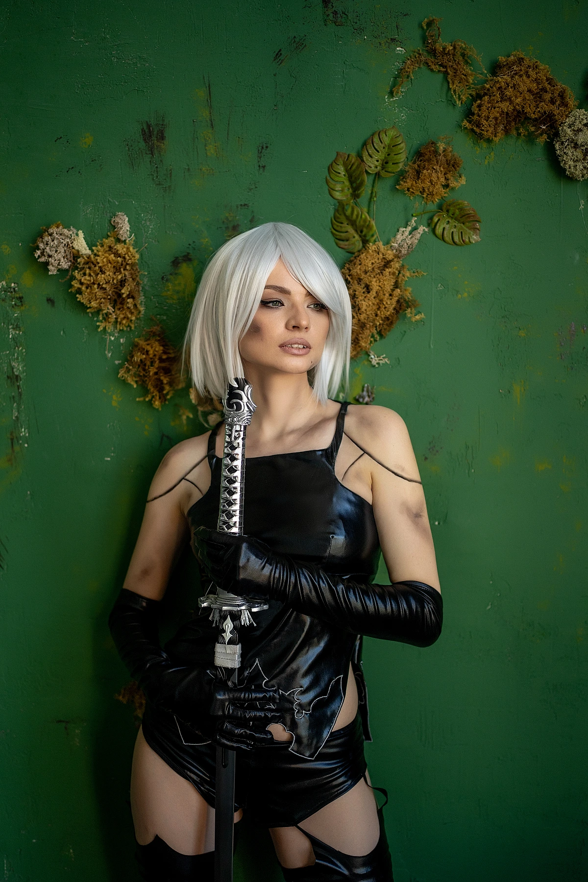 A2 NieR Automata cosplay standing portrait, vertical katana blade hold, glossy black latex outfit, white bob hair, green wall background, quiet authority pose