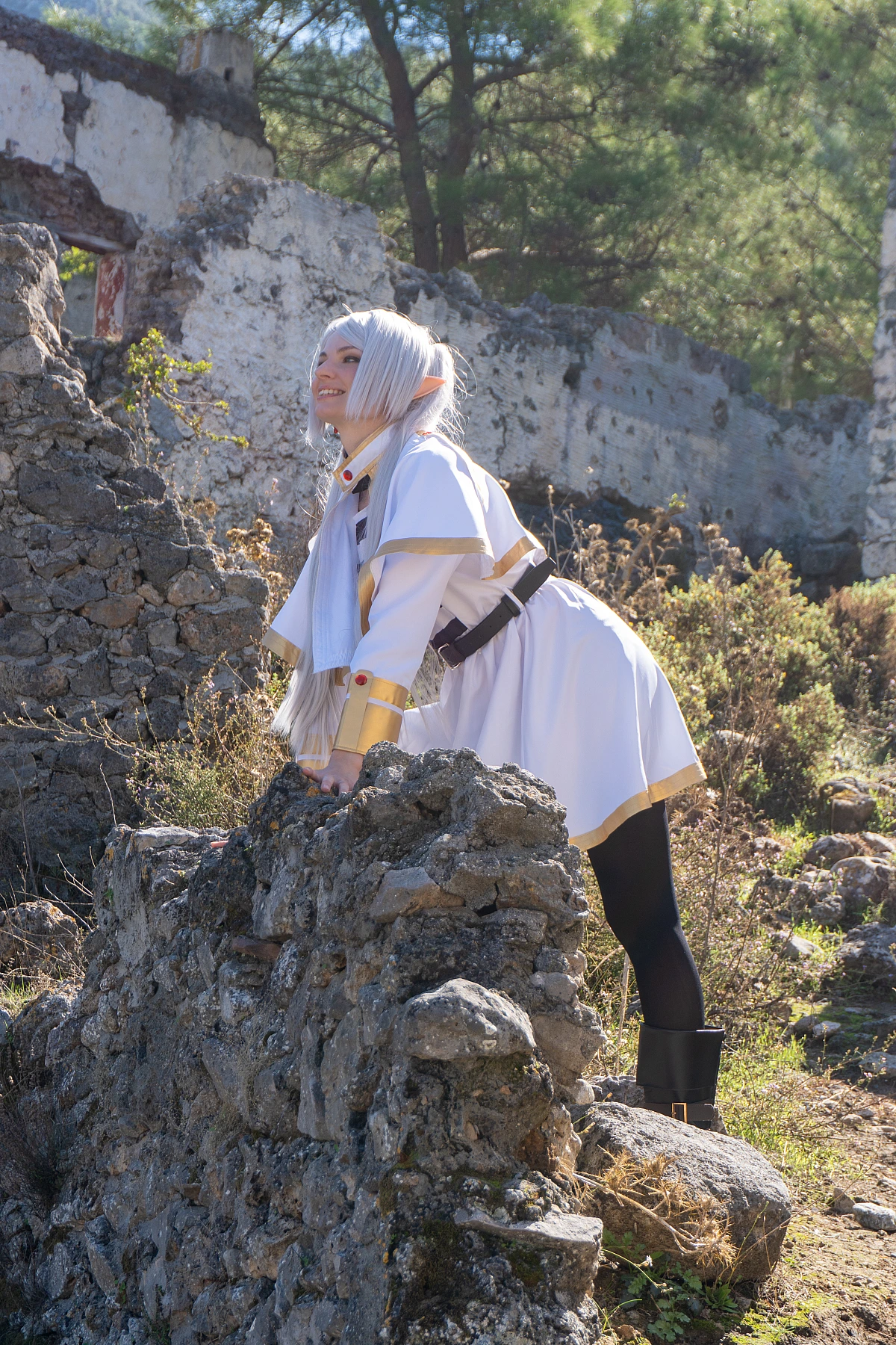 Frieren cosplay leaning low stone wall silver hair elf ears white gold mage outfit ruined architecture natural daylight gentle expression