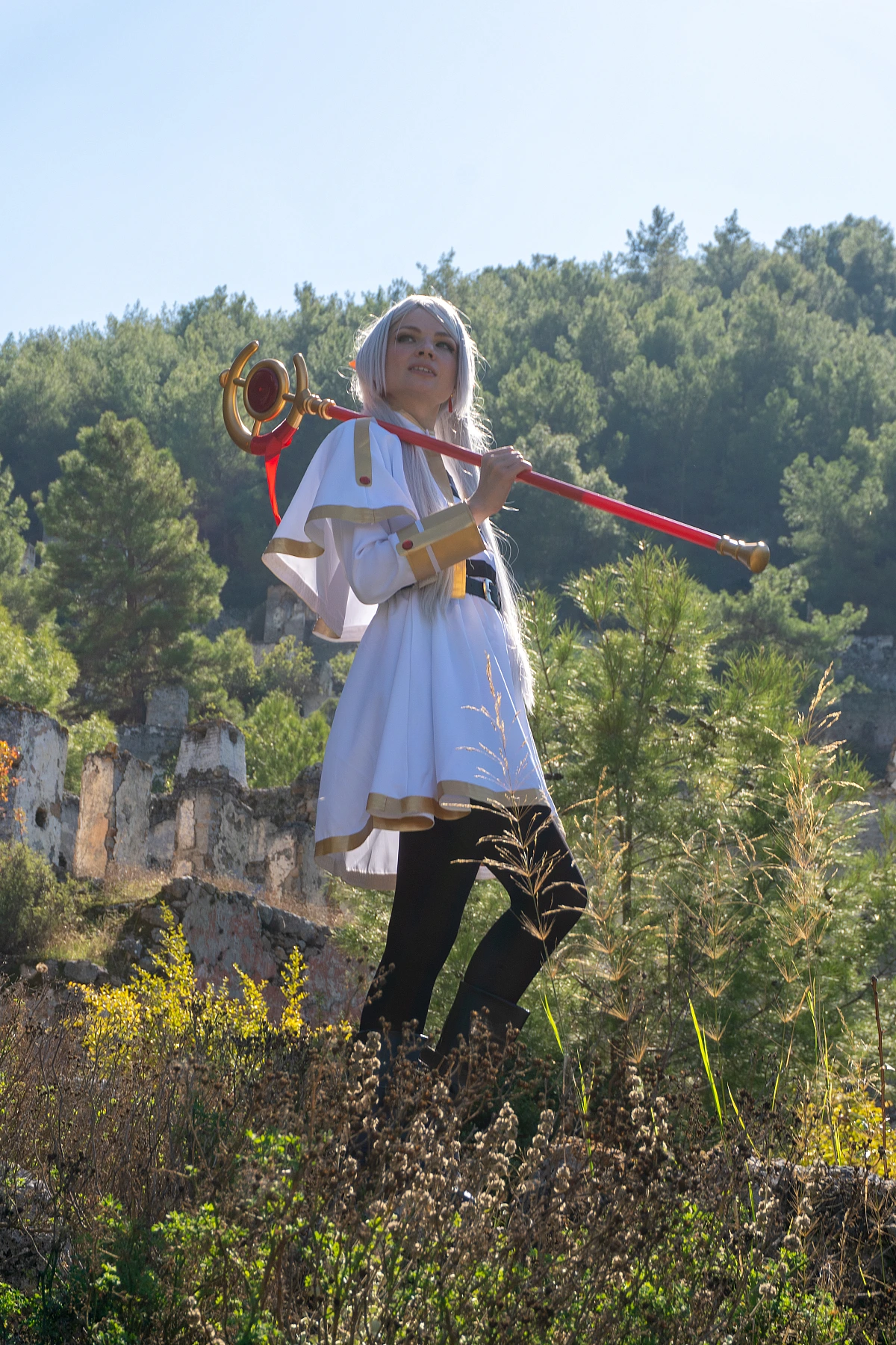 Frieren cosplay staff resting on shoulder standing tall silver hair elf ears white gold mage dress forest ruins daylight traveler