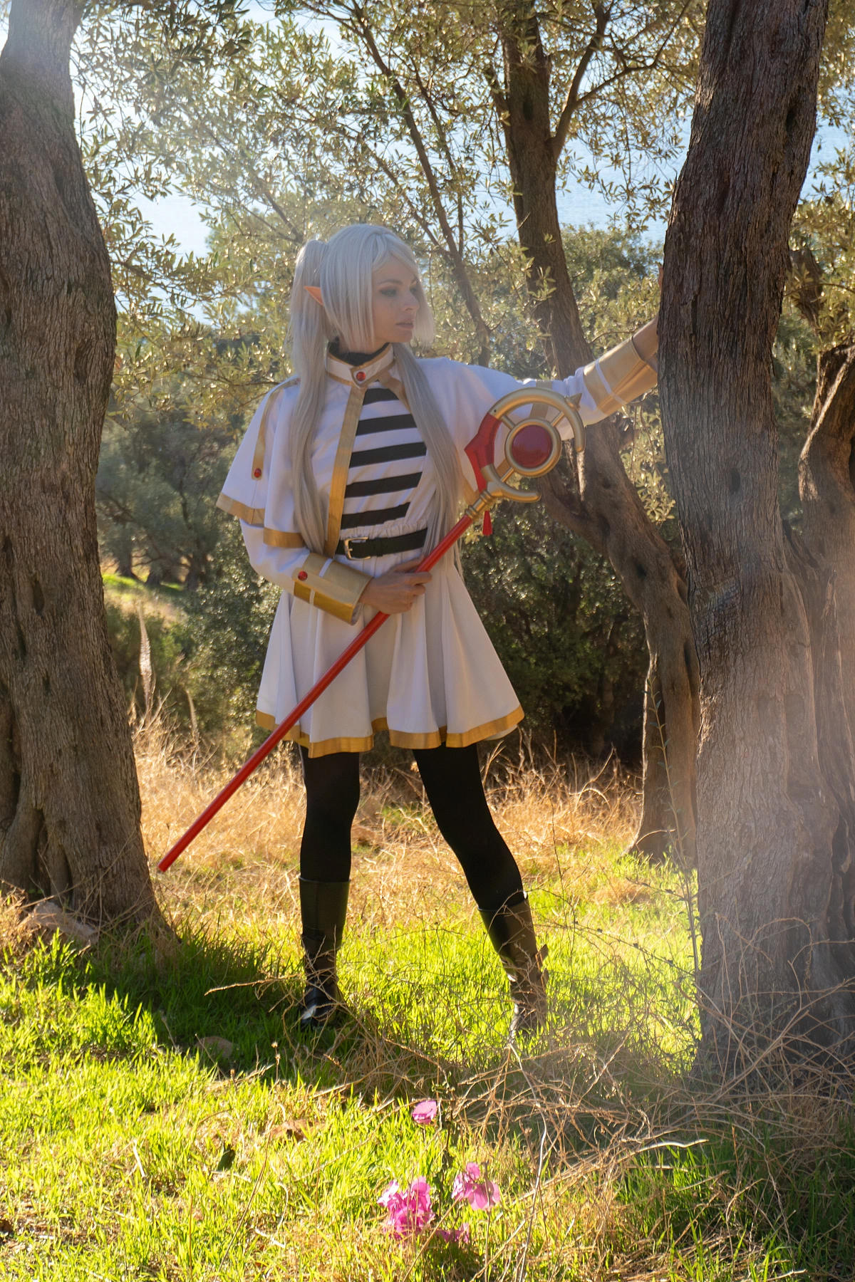 Frieren cosplay standing between two trees staff held vertically silver hair elf ears white gold mage dress forest clearing daylight