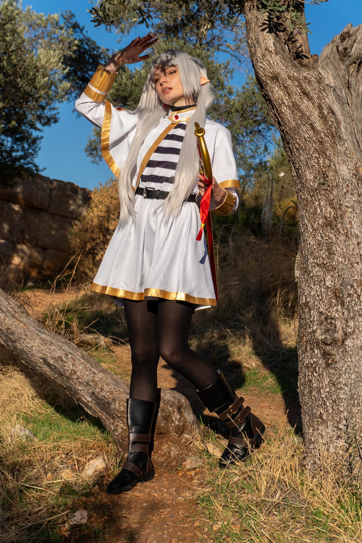 Frieren cosplay standing between trees raised hand toward sunlight silver hair elf ears white gold mage dress forest light Sousou no Frieren