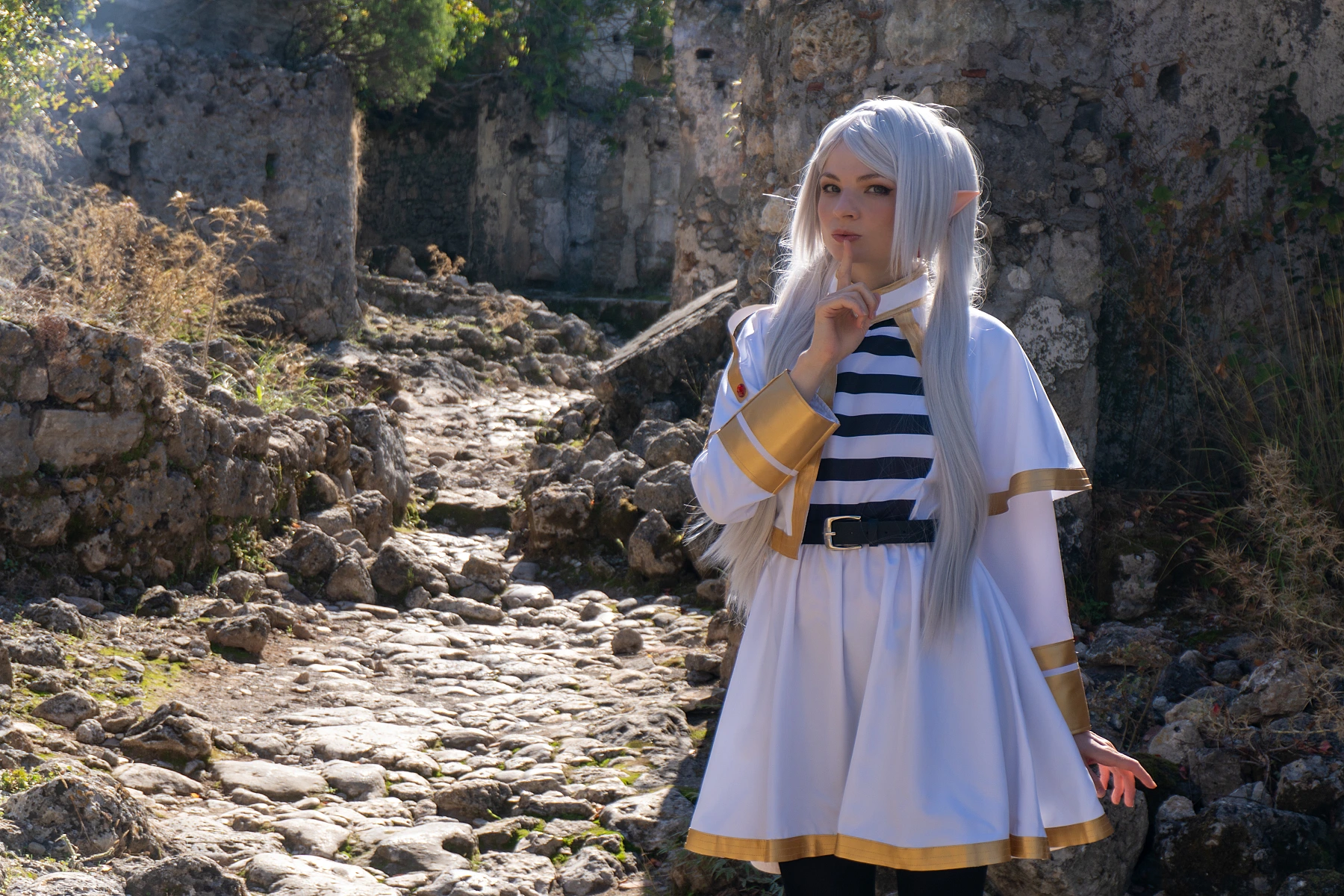 Frieren cosplay finger to lips silence gesture silver hair elf ears white gold mage dress narrow stone passage soft daylight