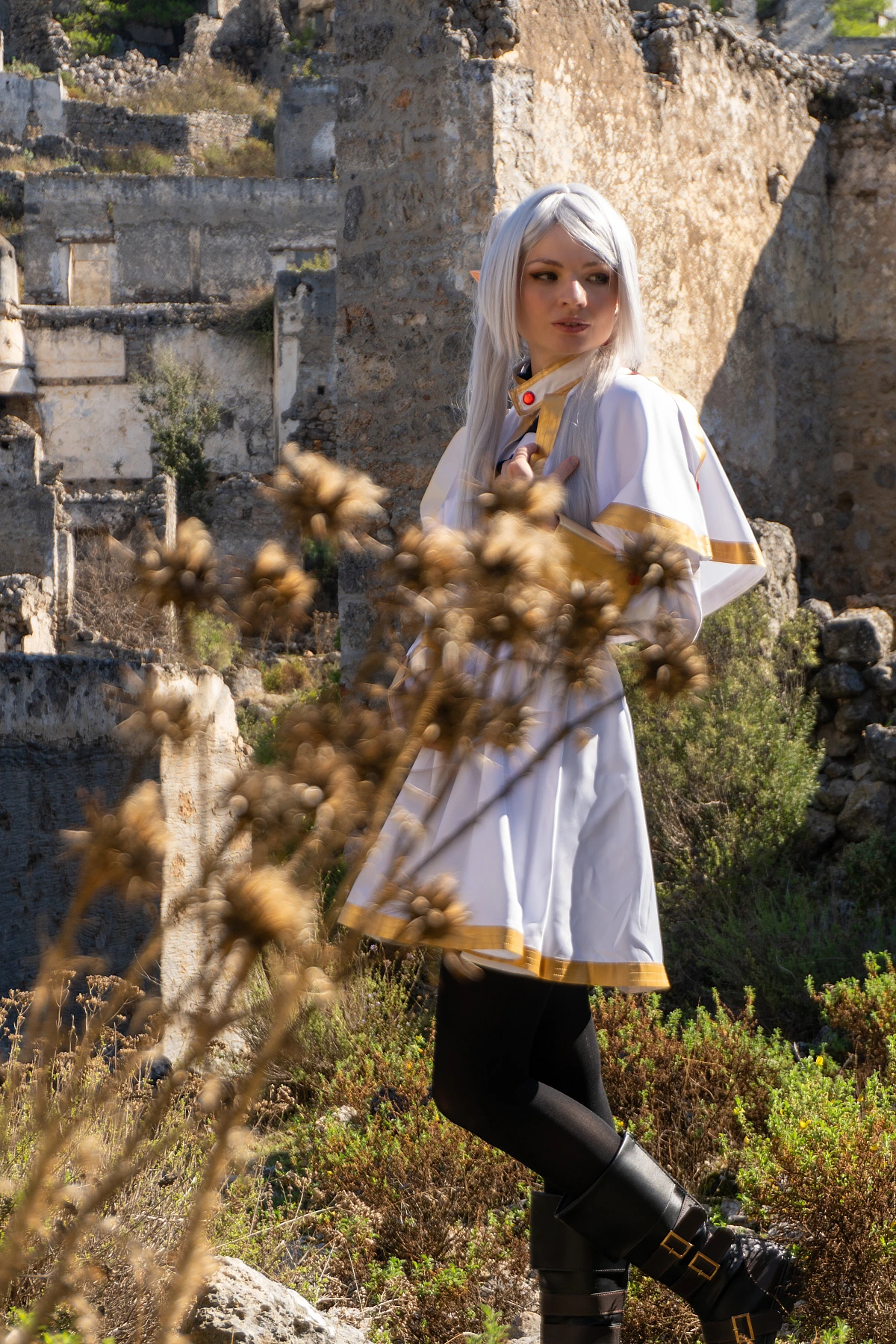 Frieren cosplay over-shoulder look hand on chest silver hair elf ears white gold outfit dry grass foreground ruins background