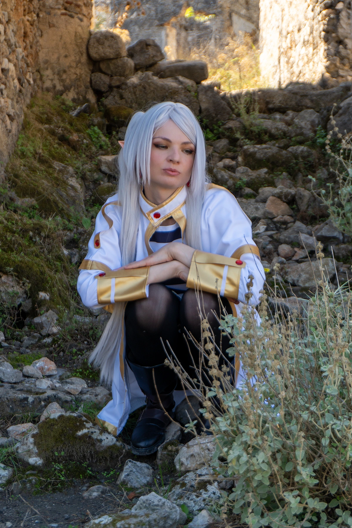 Frieren cosplay crouched stone ruins downward gaze silver hair elf ears white gold mage outfit dry grass soft daylight