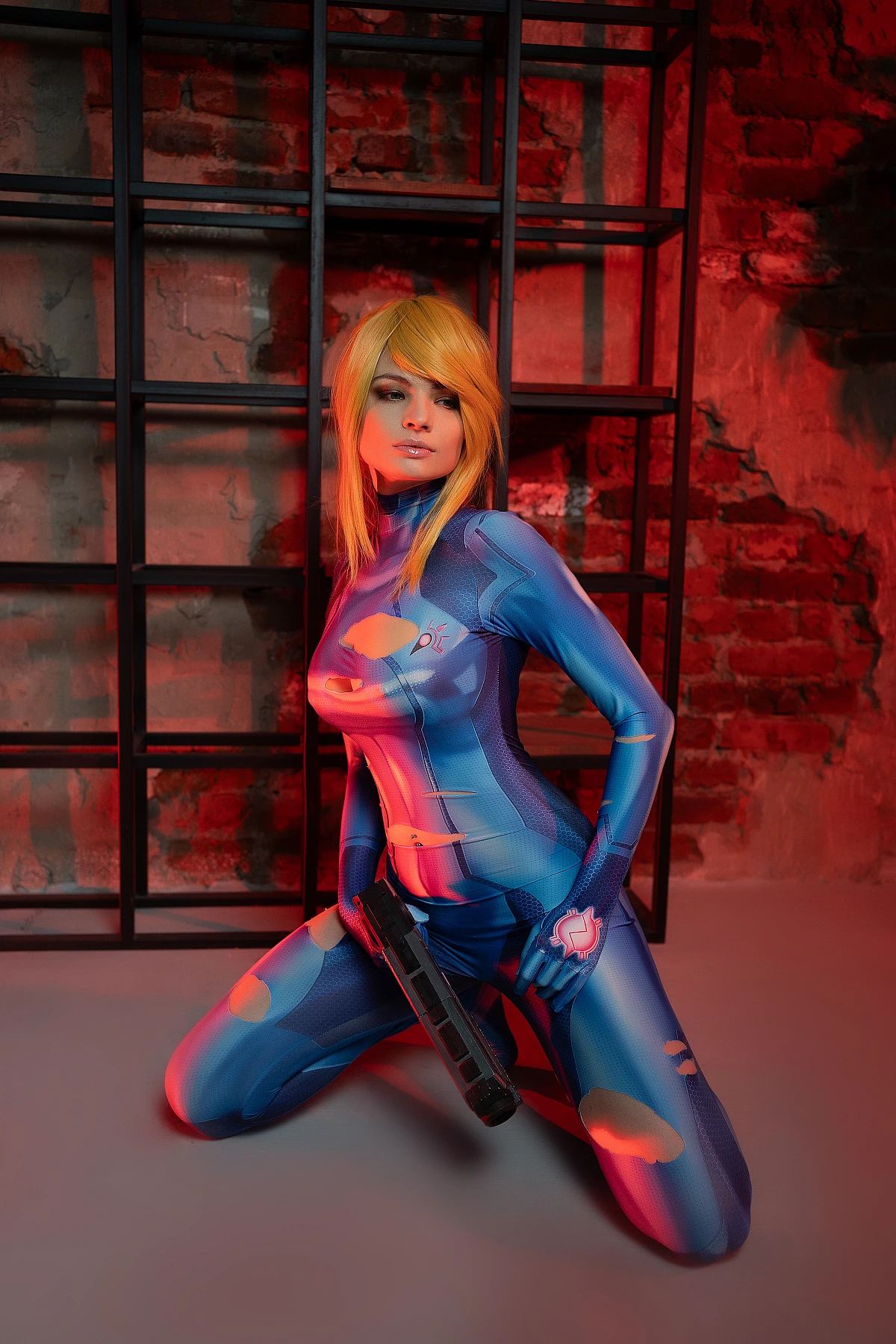 Zero Suit Samus cosplay floor-level kneeling pose, glossy blue bodysuit, sci-fi blaster, red industrial lighting, metal grid backdrop
