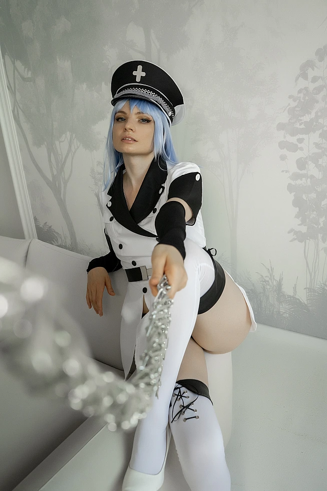 Esdeath cosplay from Akame ga Kill! seated with crossed legs on a white sofa, holding a silver chain out toward the camera