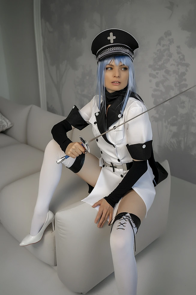 Esdeath cosplay from Akame ga Kill! sitting astride a white sofa arm, holding a blue-hilted rapier with the blade angled upward
